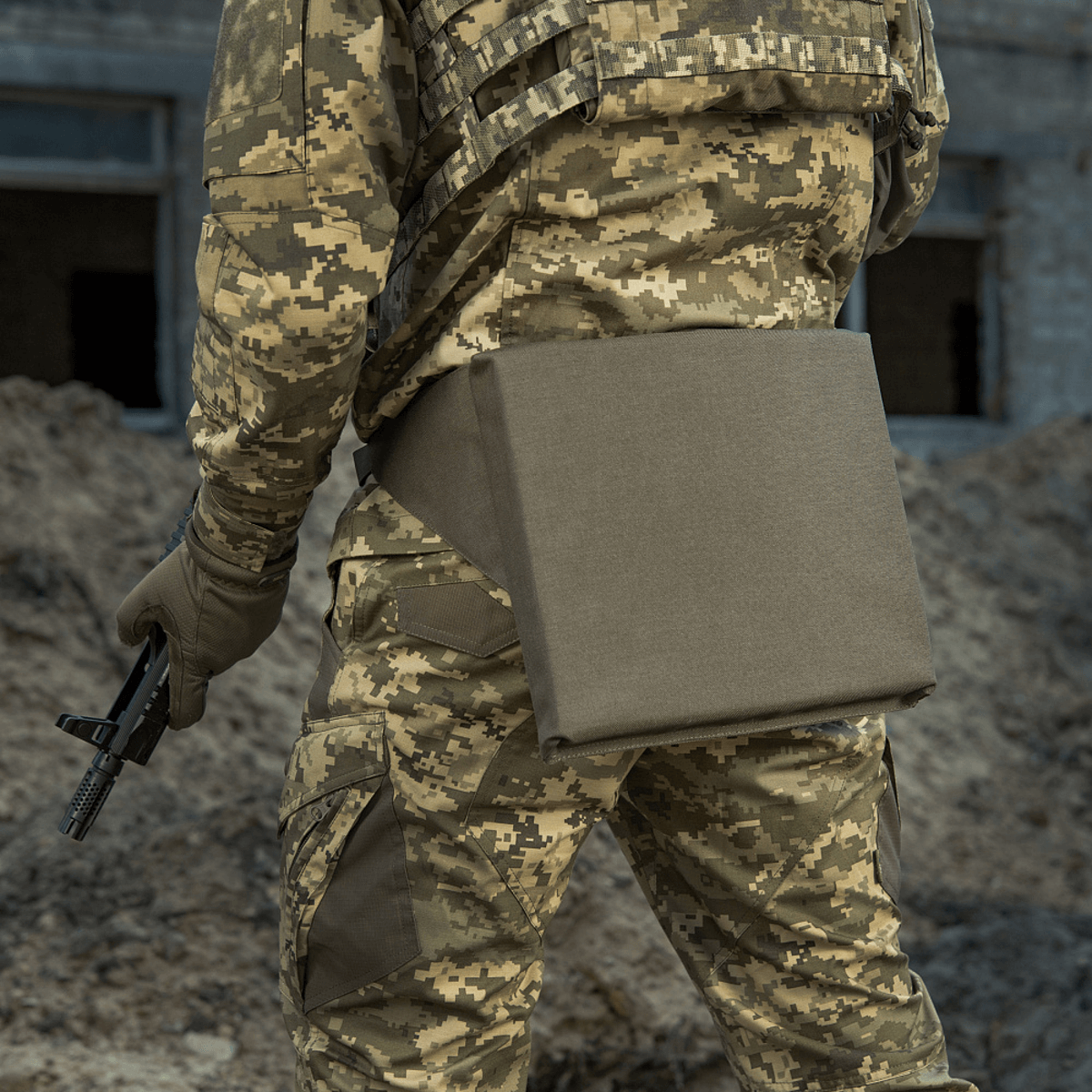 M-Tac Siddeunderlag med bælte i brug af en person i camouflagedragt, udendørs med ru terræn i baggrunden.