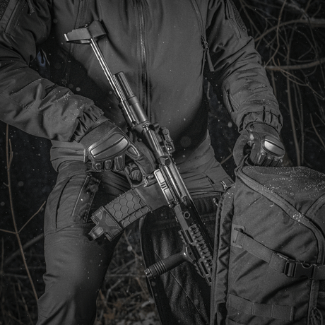 En person holder et våben ved siden af en M-Tac Rygsæk Gen.II Elite Large i en skovområde. Rygsækkens holdbare design er tydeligt.