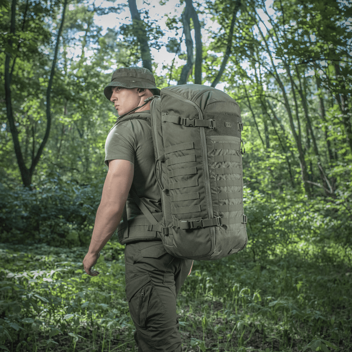 Mand med M-Tac Rygsæk Gen.II Elite Large i skovterræn, ergonomisk design med holdbar Cordura, ideel til outdoor og våbentransport.