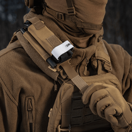 Person i bruna kläder bär M-Tac elastisk tourniquetficka Molle Gen.III fäst på västen, utrustad med taktisk utrustning.