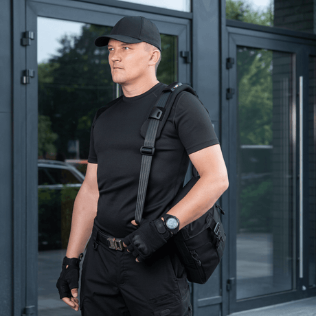 Man bär väska med M‑Tac axelremspad 50 mm Elite för ökad komfort och stöd, iklädd svart klädsel framför modern byggnad.