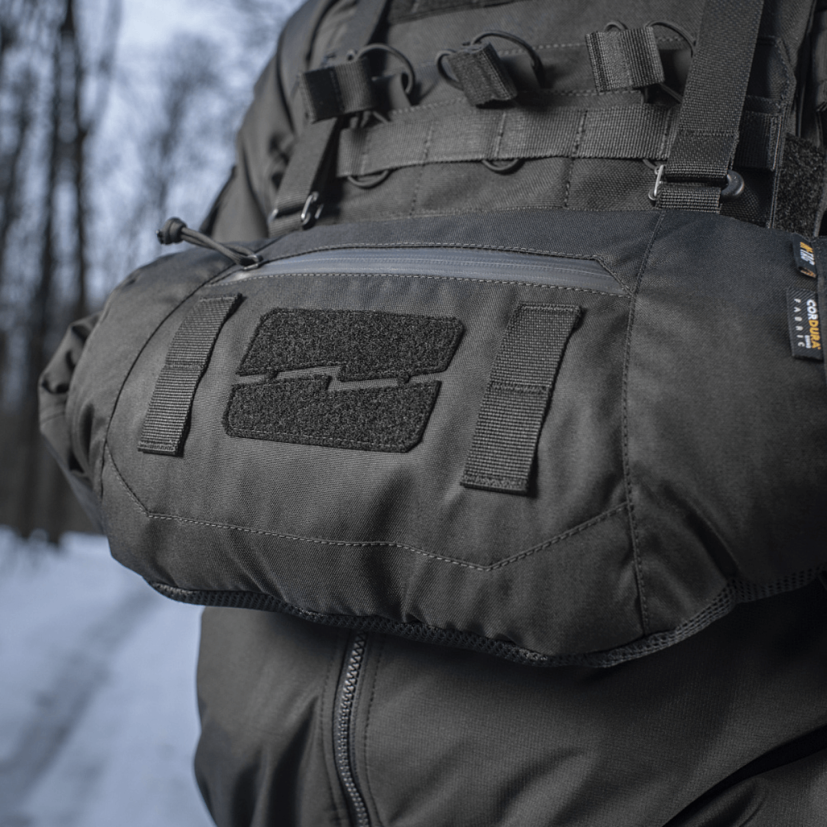 Person iført M-Tac Taktisk Håndvarmer Elite i snefyldt skovmiljø, fremstillet af slidstærkt Cordura 500D med blødt Polartec® fleecefor.