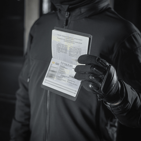 Person med M‑Tac passfodral håller ett pass, skyddar mot slitage och skador, modern polymer, stilrent design, idealiskt för resor.
