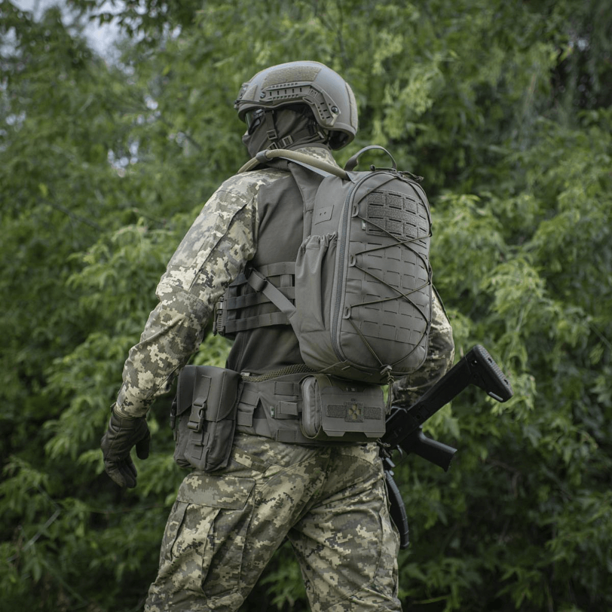 Soldat i camouflageuniform og taktisk udstyr i skovområde, bagfra