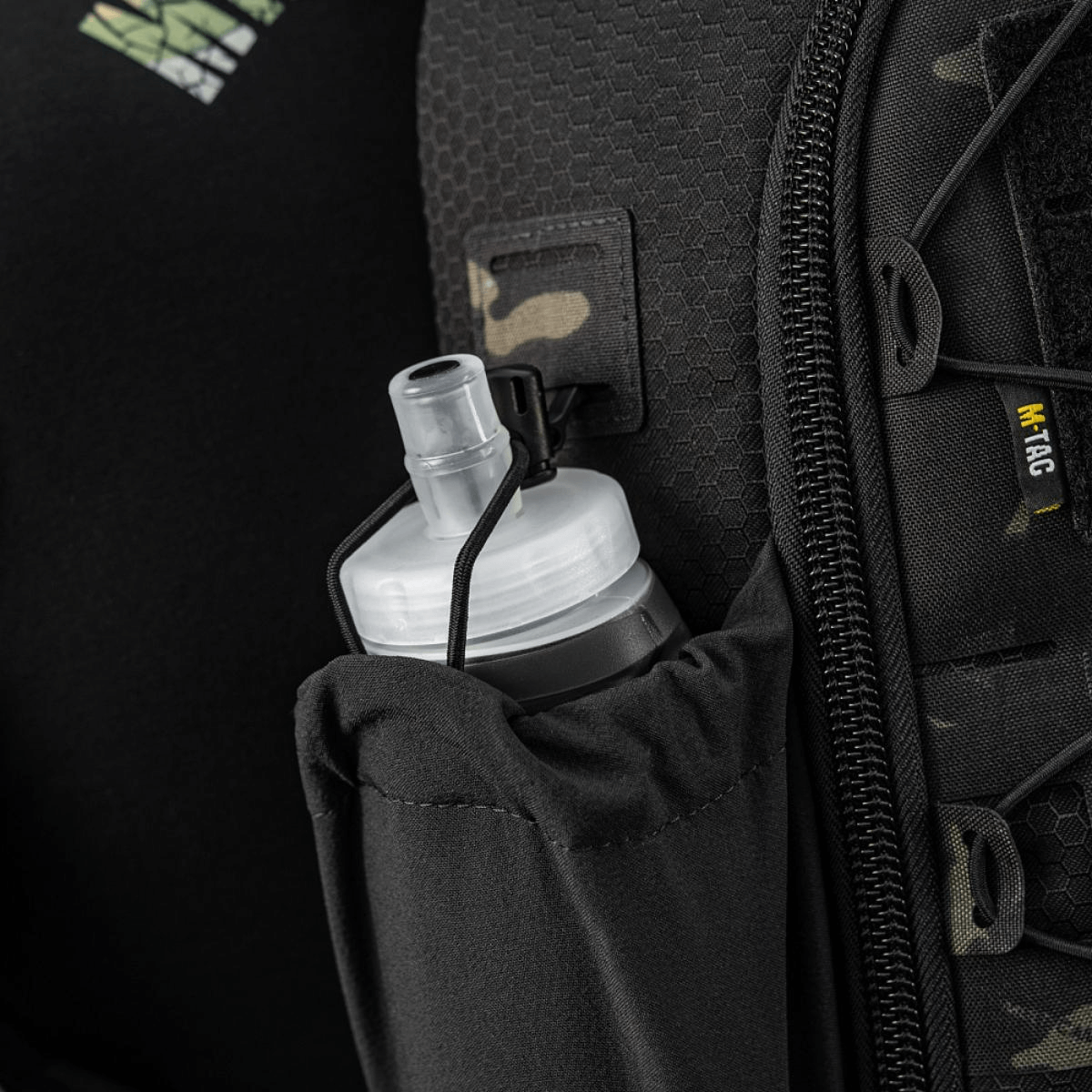 Sort rygsæk med netlomme og hvidt, rund låg til drikkeflaske, perfekt til udendørs aktiviteter. Camouflagedetaljer og lynlås synlige.