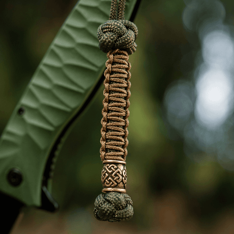 M-Tac Knife Lanyard Celtic i flätad paracord med dekorativt keltiskt mönster fäst vid greppdelen av en kniv.