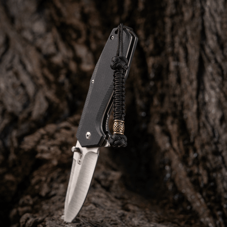 M-Tac Knife Lanyard Celtic i flätad paracord monterad på en kniv mot en rustik bakgrund.