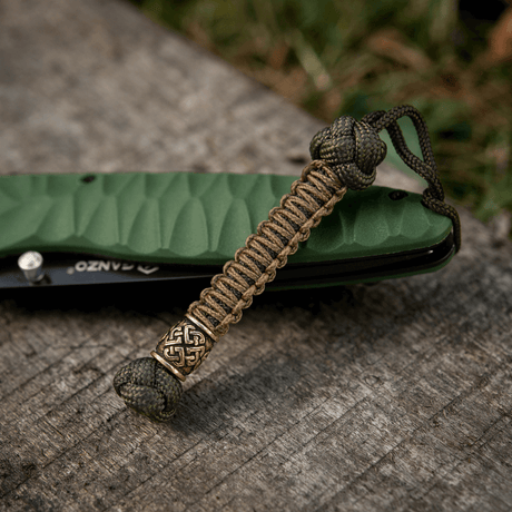 M-Tac Knife Lanyard Celtic i flätad paracord bredvid en grön fickkniv på träyta.