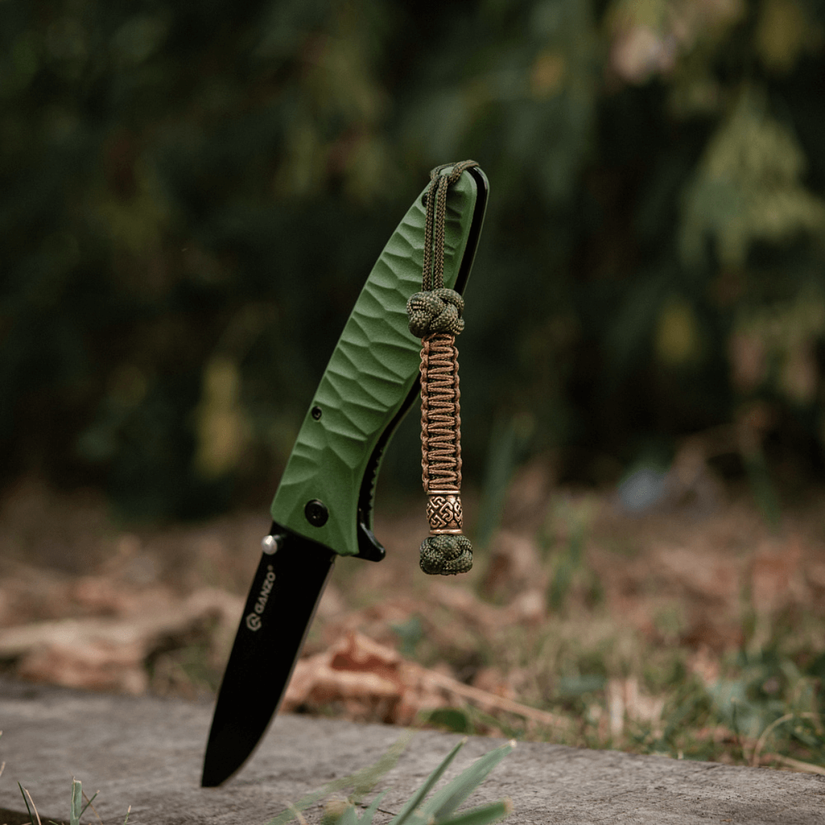 M-Tac Knife Lanyard Celtic - grøn foldekniv med flettet paracord lanyard i udendørs miljø.