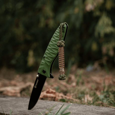 M-Tac Knife Lanyard Celtic - grön hopfällbar kniv med flätad paracord-lanyard i utomhusmiljö.