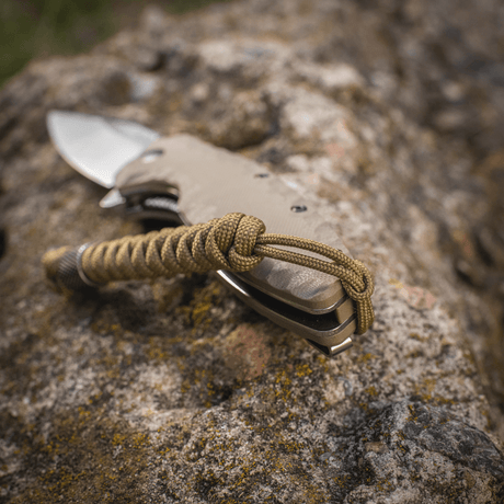 M-Tac Viper Lanyard Stainless Steel fäst vid kniv, visad på stenyta. Flexibel förlängningsdel i flätad paracord.