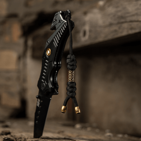 M-Tac Zeus Lanyard i flätad paracord fäst vid svart kniv på rustik yta, flexibelt tillbehör för snabb åtkomst till utrustning.