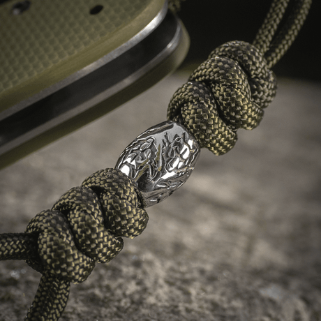 M-Tac Zeus Stainless Steel Lanyard med flätad paracord och dekorativ metallpärla på en kniv, perfekt för snabb åtkomst.