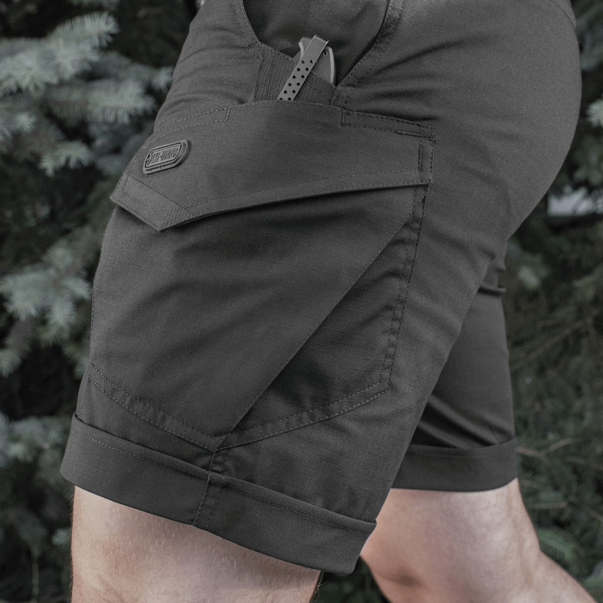 Herre iført M-Tac Shorts Aggressor Short, sorte knælange shorts med mange lommer, ideel til taktisk brug i varmt vejr. Lavet af polycotton flex rip-stop.