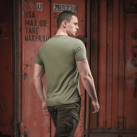 Person iført armygrøn M-Tac T-shirt Viking med ryggen til, gående forbi container i bymiljø.
