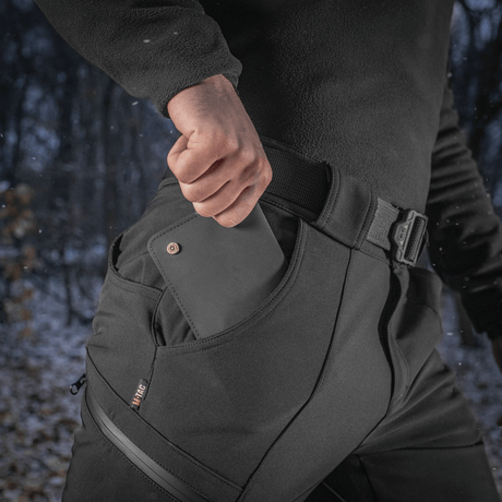 M-Tac Bukser Soft Shell Vinter i sort, med fleeceforing, viser en mand der putter en smartphone i lommen, ideel til kuldeklimaer.