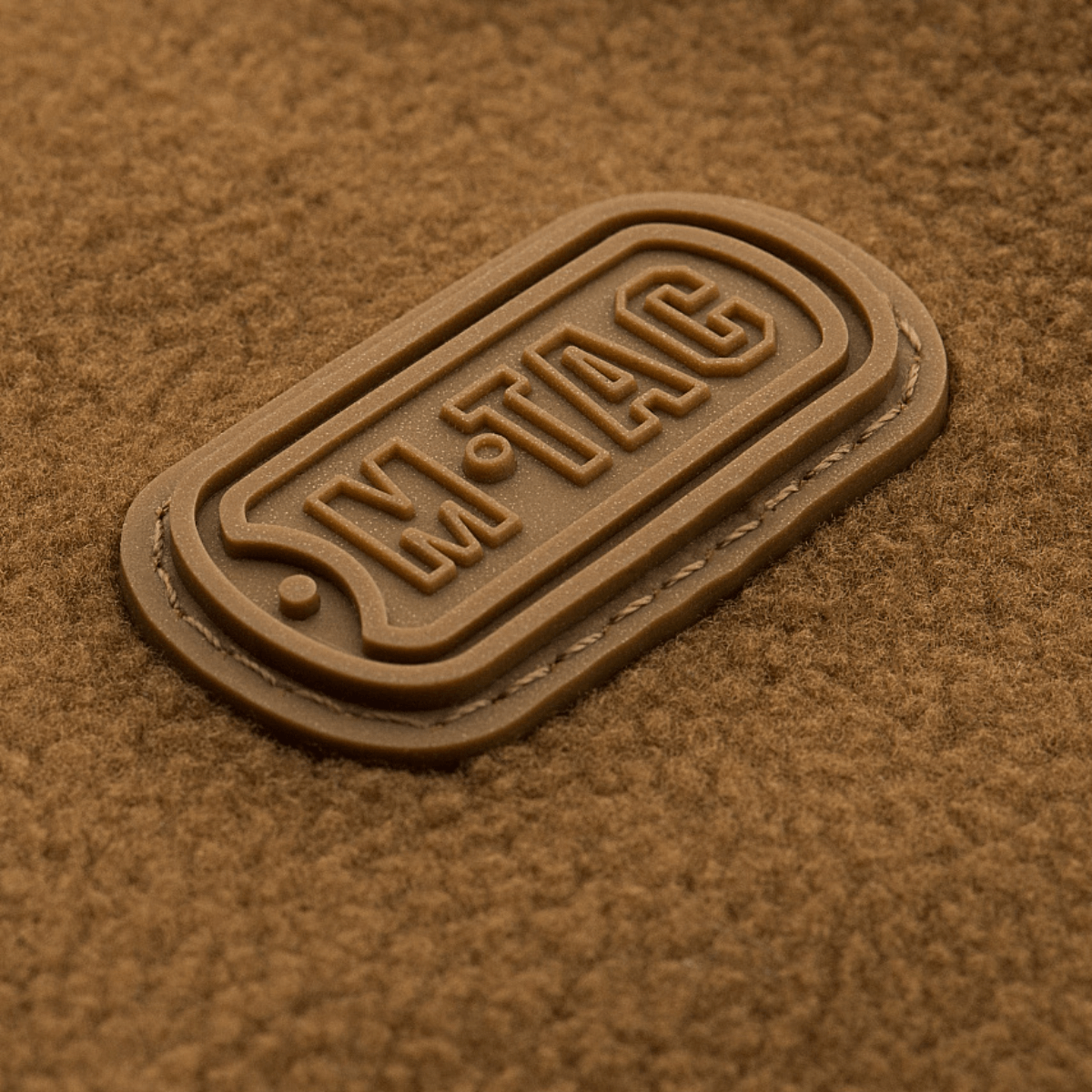 M-Tac Alpha Microfleece Jakke Gen.II logo på brun fleece materiale, der viser jakens styrke og holdbarhed til taktisk brug.