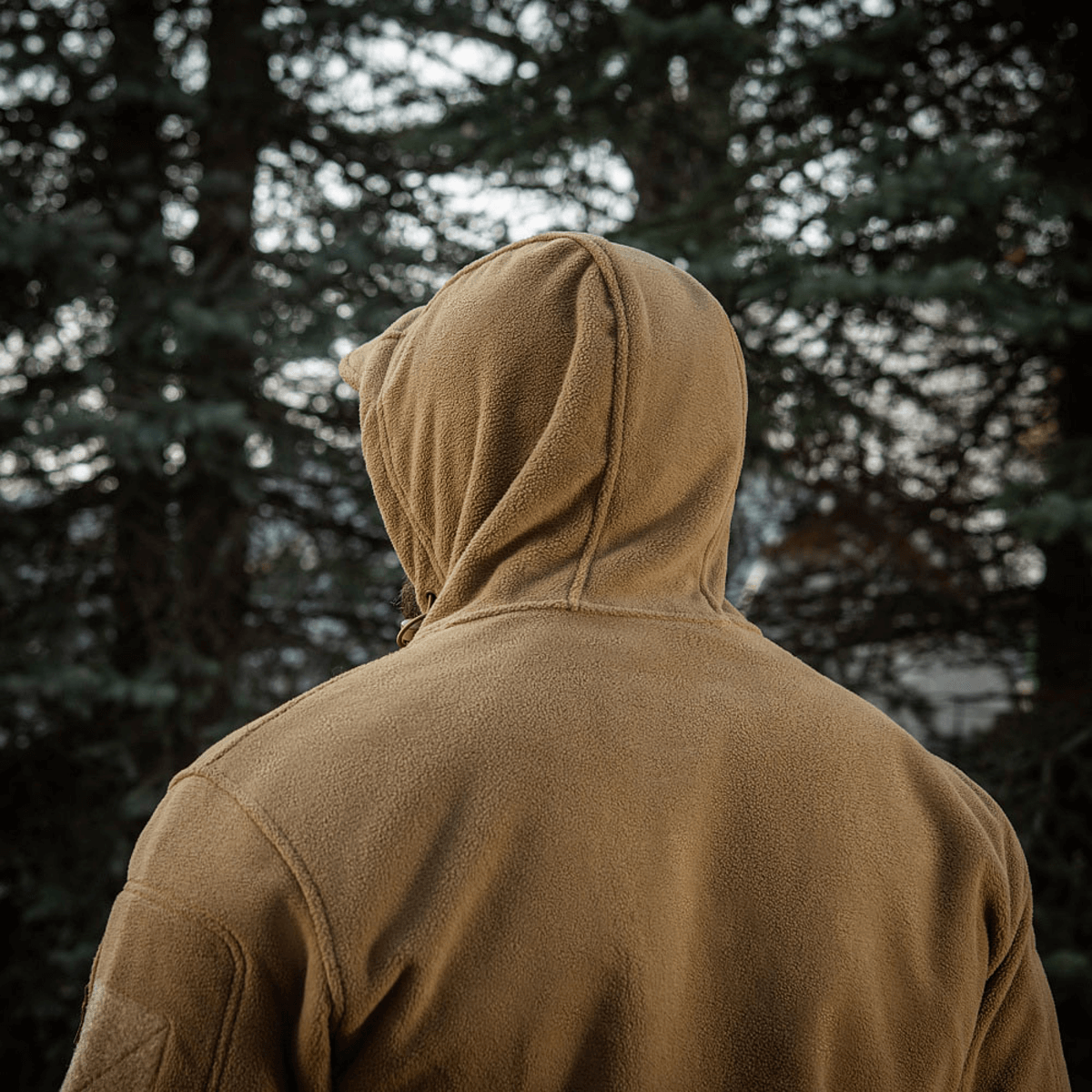 Person iført M-Tac Division Gen.II Windblock Fleecejakke i et skovområde, set bagfra, fremhæver jakken til koldt vejr.