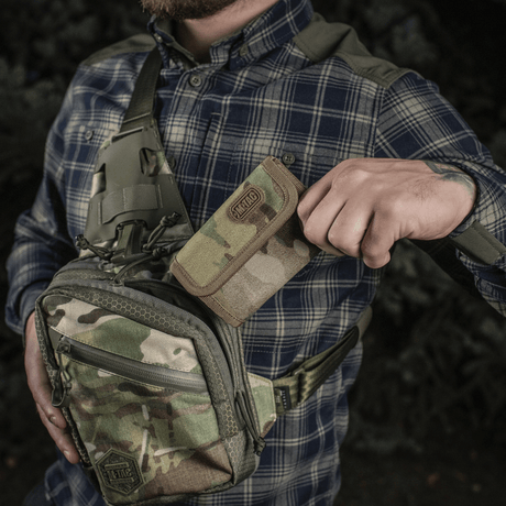 Man håller M-Tac Plånbok Elite Gen.II i Cordura 500D bredvid en kamouflage-ryggsäck, utomhusmiljö.