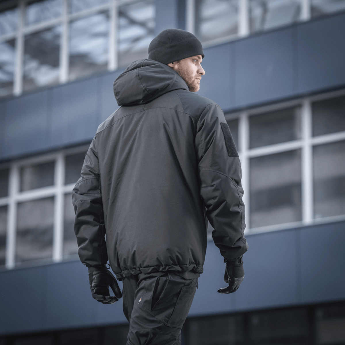 Mand bærer M-Tac Alpha Gen.III Pro Vinterjakke, perfekt til temperaturer ned til -20° C, foran moderne bygning.