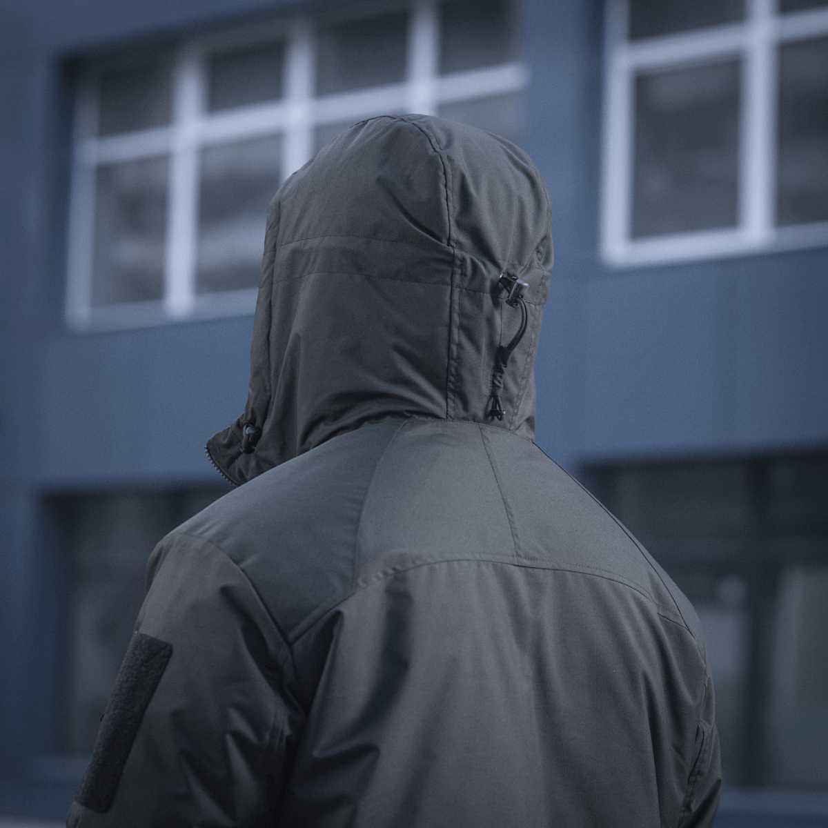 M-Tac Alpha Gen.III Pro Vinterjakke set bagfra i bymiljø, viser justerbar hætte og forstærkede områder på skuldre