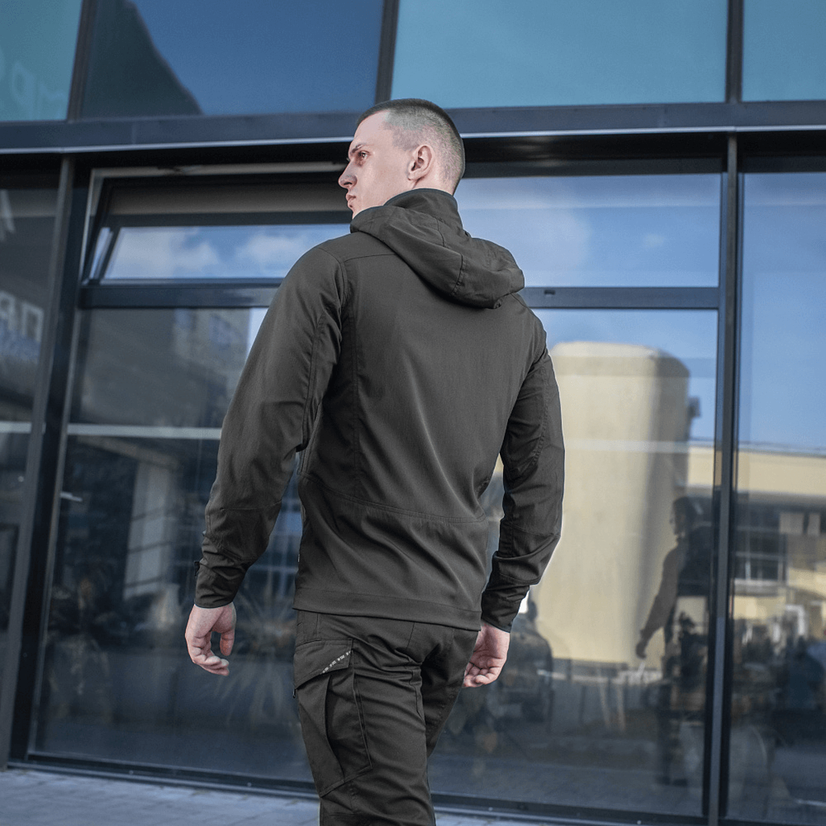 Mand iført M-Tac Flash Jakke foran en moderne bygning, ideel til både hverdag og sport, uden velcrofelter på ærmerne.