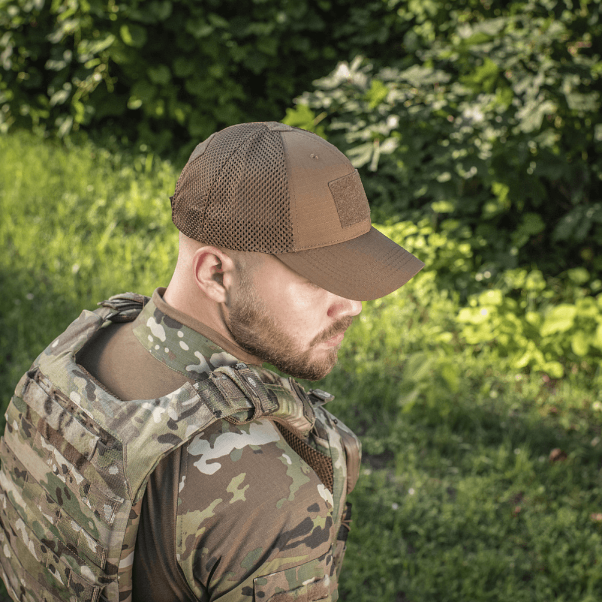 M-Tac Baseball Cap Ripstop Elite Flex i brun bæres af en person i camouflagetøj på en solrig dag omgivet af grønne buske og græs.