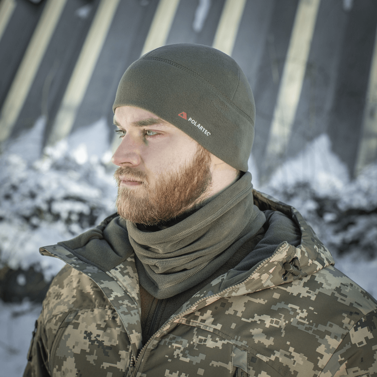 Mand iført grå M-Tac Fleece Watch Cap Light Polartec hue i sneklædt landskab, bæret med camouflagetøj, perfekt til vintervejr.