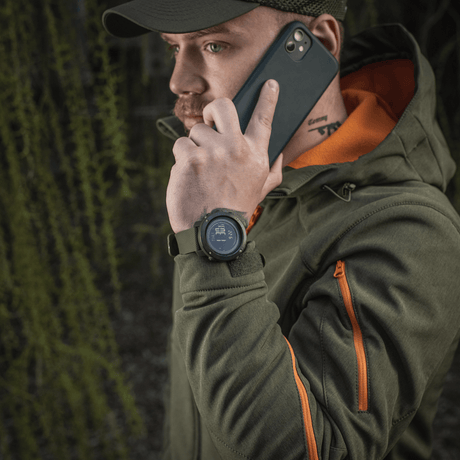 Man som bär M‑Tac Multifunktionellt Taktiskt ur medan han pratar i telefon, skog i bakgrunden