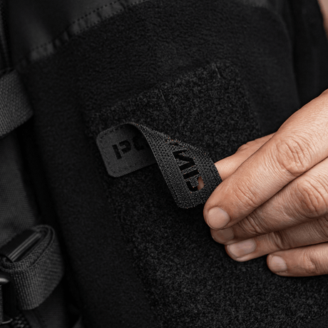 Person fäster M‑Tac Patch Poland End-to-End 25x80 på kläder med velcro, visar detalj av laserskuren inskription och hållbart Cordura 500D-material.