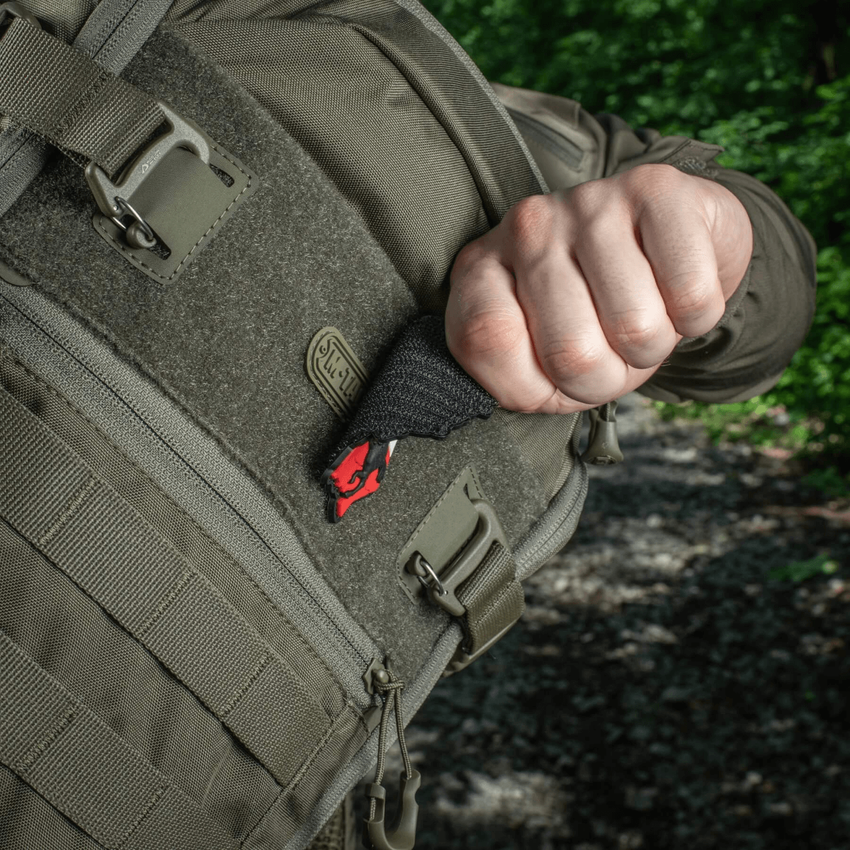 Person trækker en M-Tac Patch Husaria 3D PVC morale patch ud af en taktisk rygsæk, viser vingede husar design mod grøn skov baggrund.