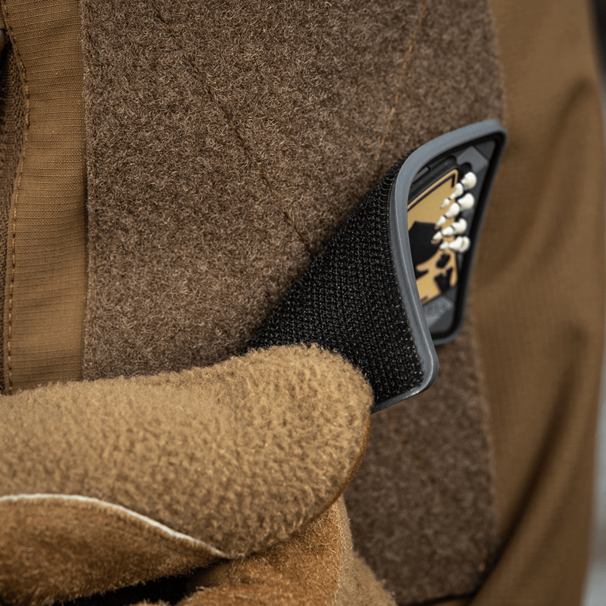 Brun handske fastgør M-Tac Patch Joker Skull 3D PVC på jakke med velcro.