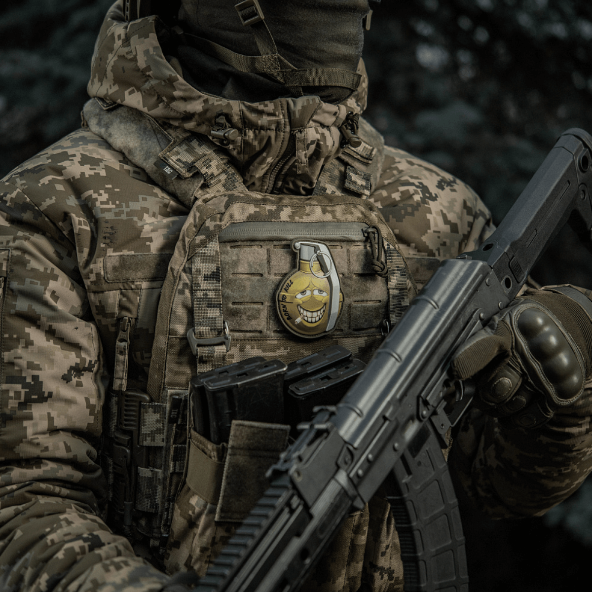 Soldat med camoufleret jakke bærer M-Tac Patch M67 Granat 3D PVC på vesten, humoristisk emoji-design, taktisk militærudstyr.