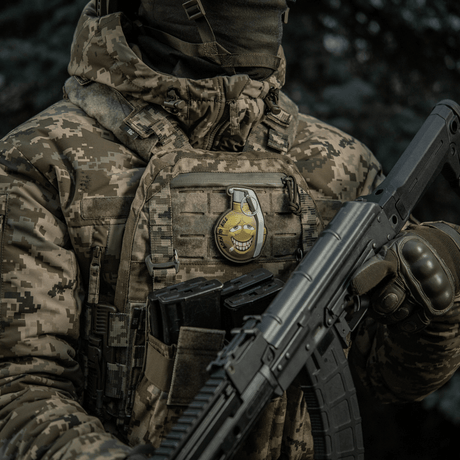 Soldat i kamouflerad jacka bär M-Tac Patch M67 granat 3D PVC på västen, humoristisk emoji-design, taktisk militärutrustning.
