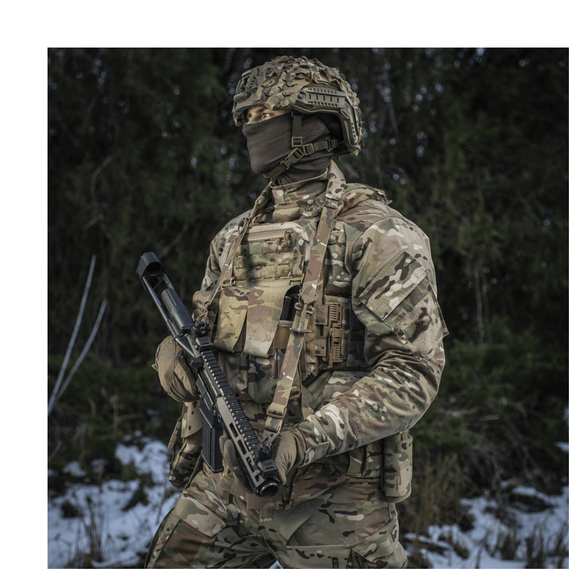 Soldat iført camouflageuniform udstyret med gevær og taktisk vest i skovmiljø.