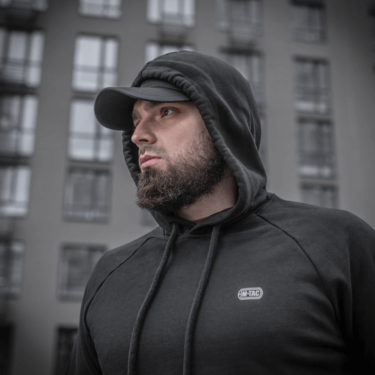 Mand iført M-Tac Cotton Raglan Hard Hoodie, 100% bomuld, med fleecefor, stilfuld til hverdagsbrug, set foran bybygning.