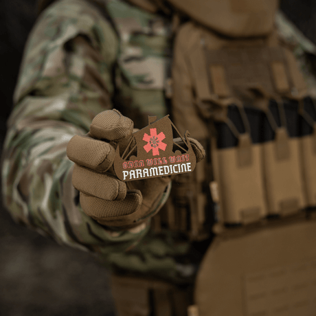 M-Tac Patch Viking Evac i en soldats hand, visar Odin Will Wait Paramedicine-motiv. Kamouflagejacka och taktisk väst i bakgrunden.