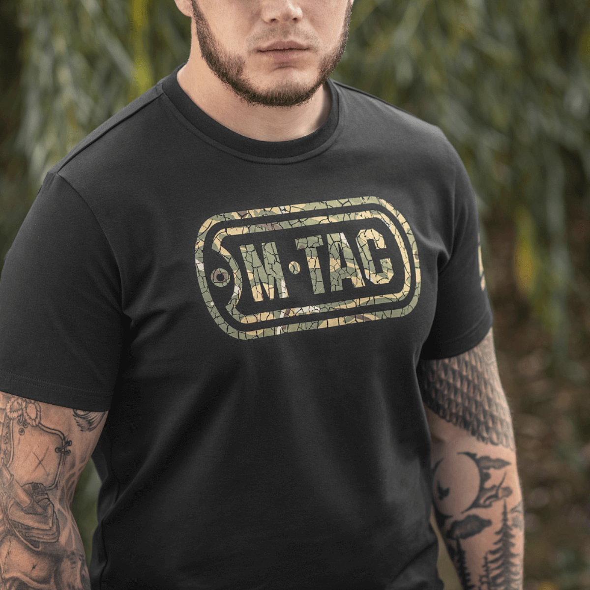 Mand iført M-Tac T-Shirt Logo i camouflage med slogan på ærmet og "Military&Tactical Since 2014" på ryggen, omringet af grønne planter.