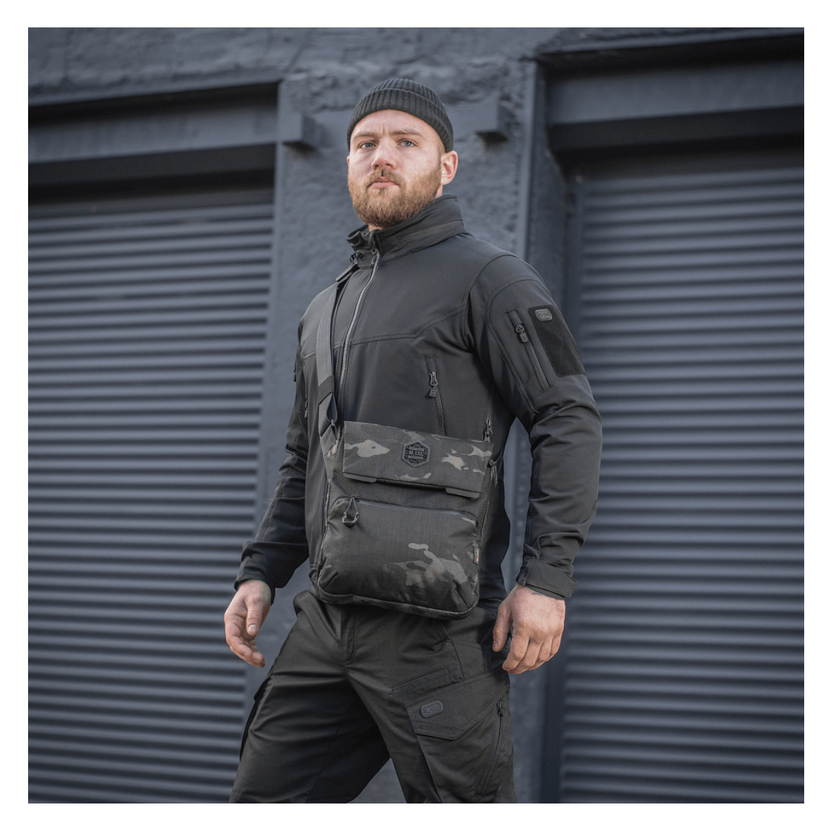 Mand iført M-Tac Konvert Taske Elite i sort Camouflage-stil foran industrielle garagedøre