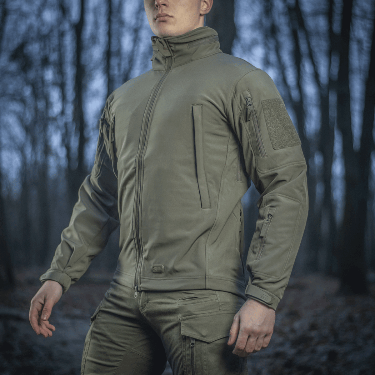 Mand iført M-Tac Soft Shell Jakke i skovmiljø, designet til at beskytte mod dårligt vejr med indre polyuretan-membran og fleece-lag.