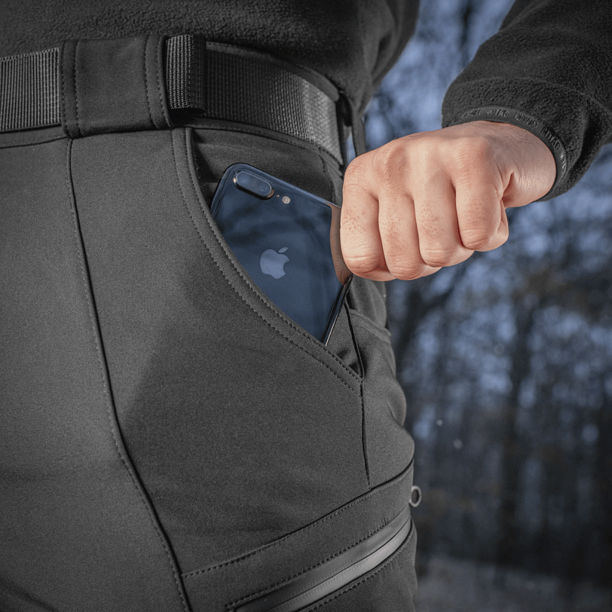 Person iført M-Tac Bukser Soft Shell Vinter med en telefon i lommen, fremhæver deres praktiske og funktionelle design til kolde klimaer.