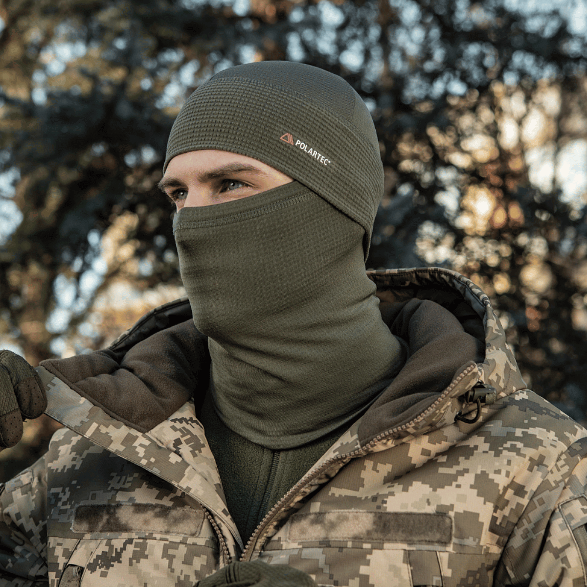 Person iført M-Tac Tube Scarf Polartec Power Grid i skovmiljø, beskyttende mod vind og kulde, multifunktionelt som halsrør og ansigtsmaske.