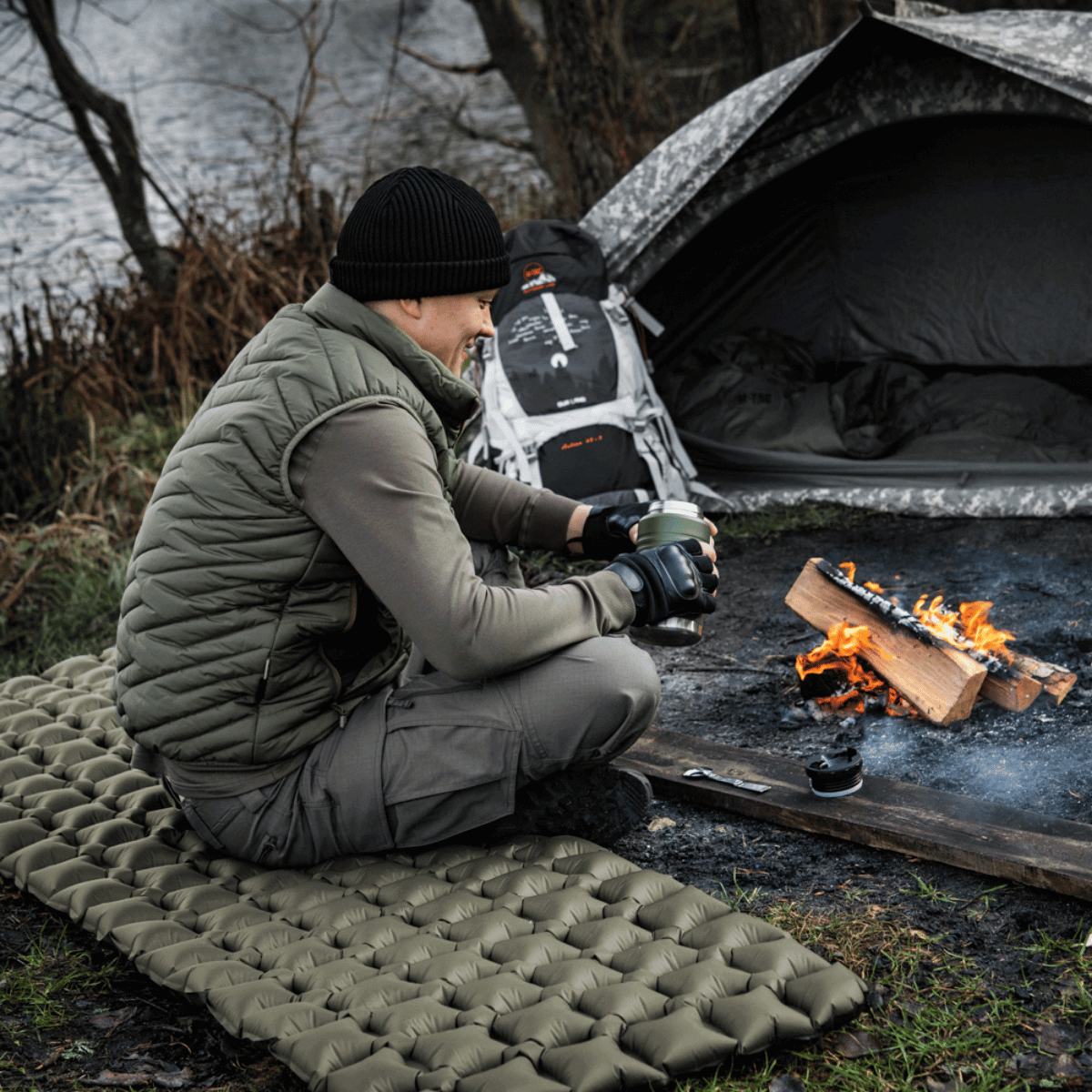 Person sidder ved bål og nyder M-Tac Oppustelig Liggeunderlag 195x60 ved en sø med telt i baggrunden, ideel til camping.