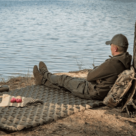 Mand hviler på M-Tac Oppusteligt Liggeunderlag 195x120 ved søbredden, omgivet af natur og en rygsæk bagved, der viser outdoor-nødvendigheder.
