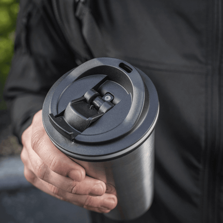Person håller M-Tac Termokopp 450 ml med ventil, designad för att hålla drycken varm eller kall, med praktisk pip och konstruktion i rostfritt stål.