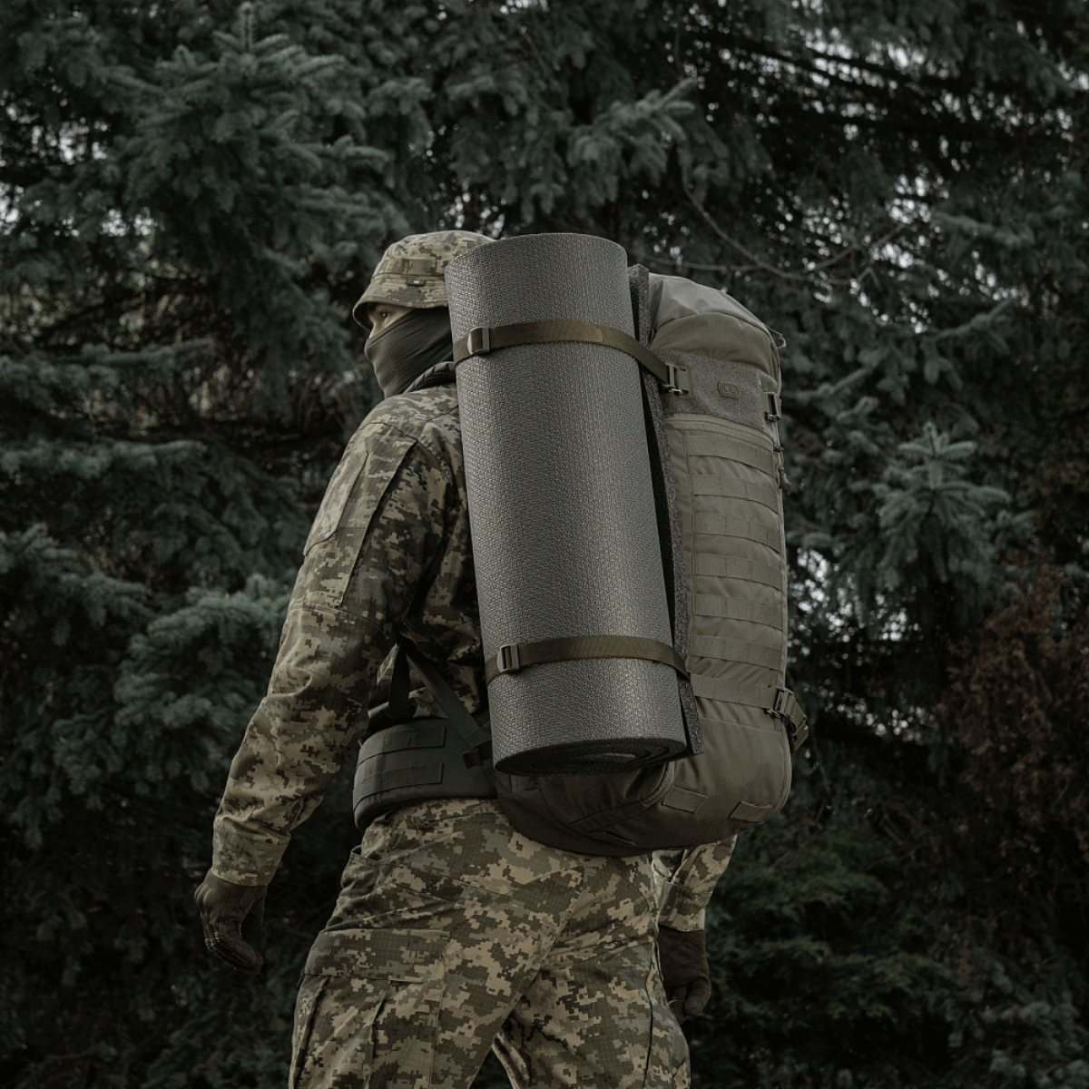 Person med camouflage, der bærer rygsæk med M-Tac Liggeunderlag 20 mm (60x180 cm) i skovmiljø.