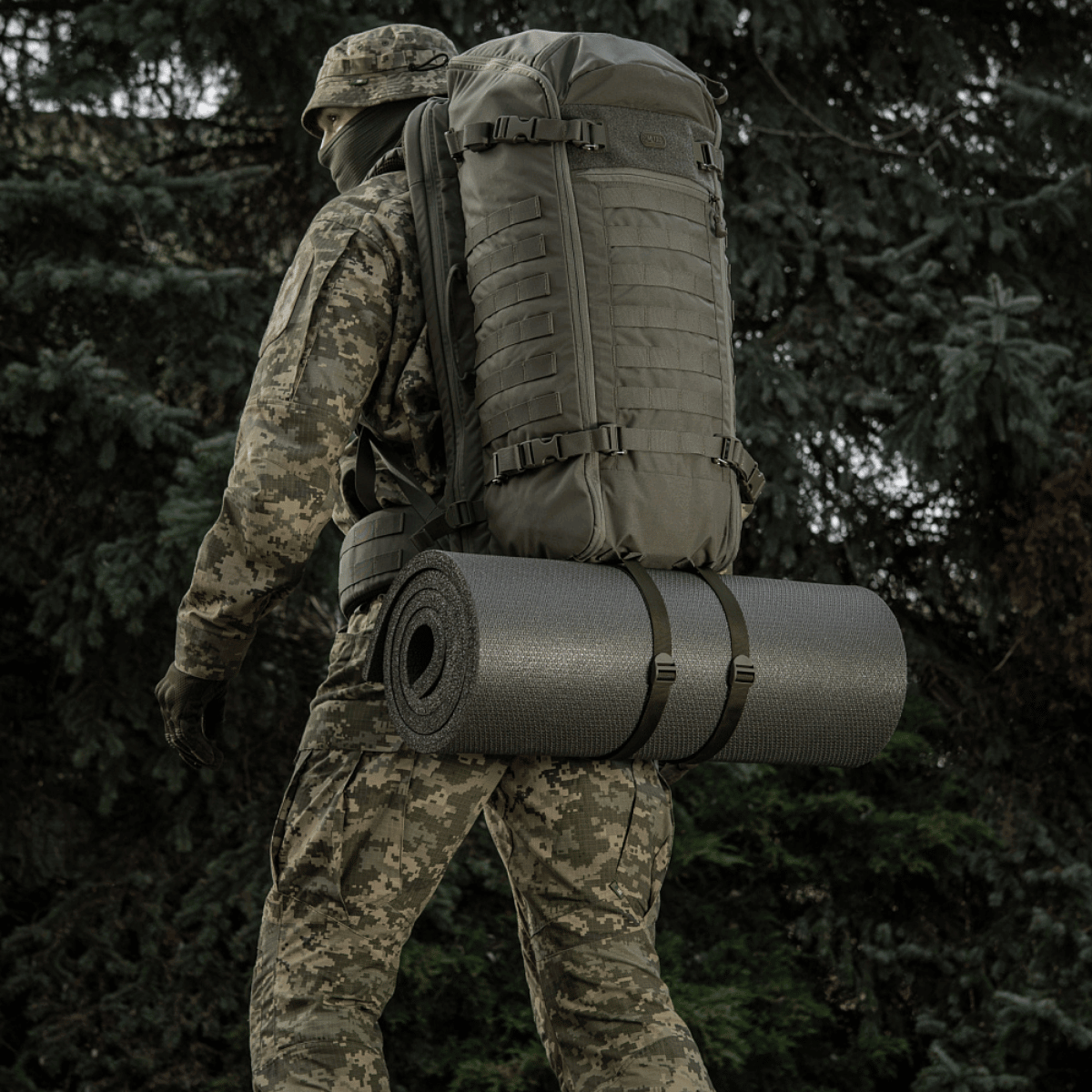 En mand iført camouflagetøj med M-Tac Liggeunderlag 20 mm (60x180 cm) fastgjort til en rygsæk i skovmiljø.