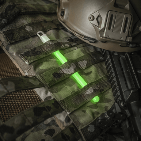 M-Tac Glow Stick 15 cm placerad på militärutrustning, giftfri och mångsidig ljusstav för säkerhets- och nödsituationer.