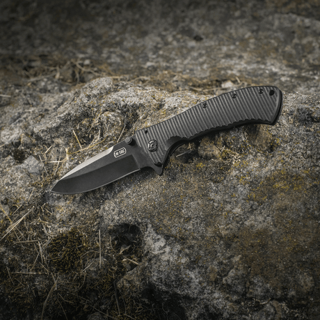 M-Tac Outdoor & Militär fällkniv - Type 7 med svart beläggning mot klippbakgrund.