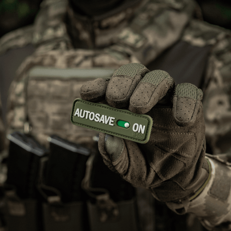 Taktisk operatör håller M-Tac Patch Autosave PVC 3D-morale-patch med velcro, lämplig för kläder eller utrustning.